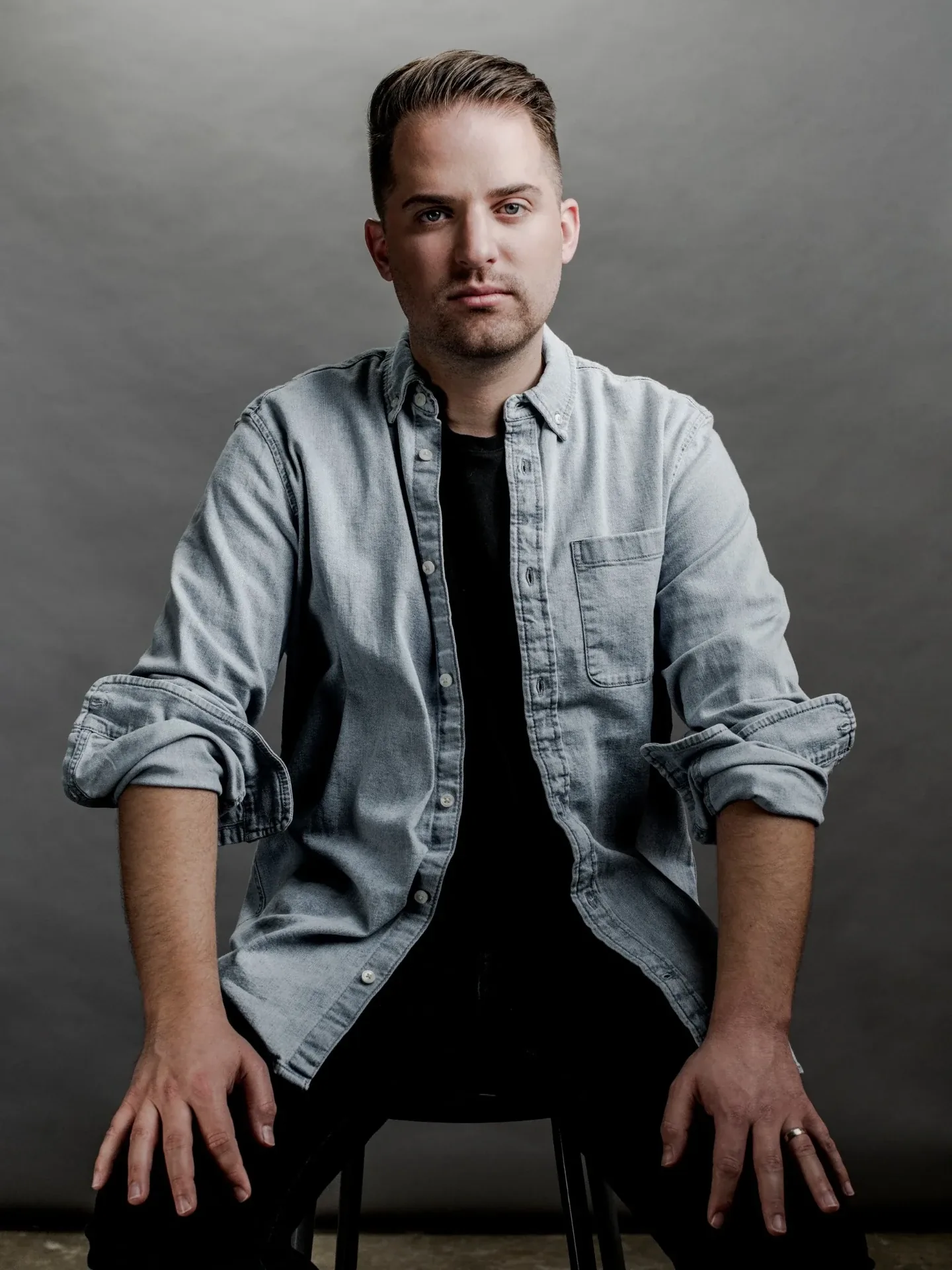 Composer Josh Tackett in a light denim shirt, seated against a grey studio backdrop